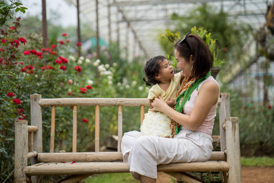 Side view of mother and daughter sitting on bench by plants