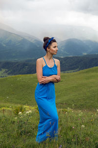 Woman in a blue long dress stands on top of a mountain in the summer on a green meadow