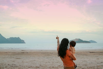 Rear view of woman holding baby girl while standing at beach against sky