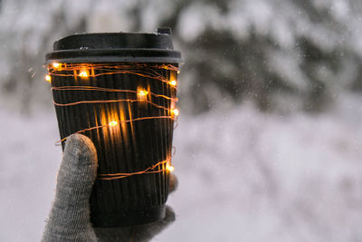 Female hand in winter warm gloves holding black eco paper cup with garland shiny light. creative 