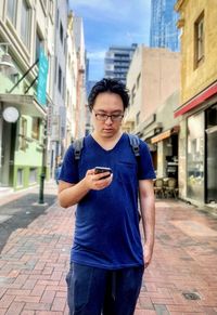 Portrait of young asian man using smartphone while standing in laneway against buildings in city.