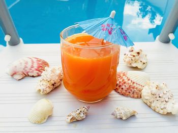 Glass of orange juice with blue umbrella and seashells near the blue pool water