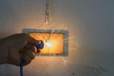 Cropped hand of person holding electric plug by sparkling outlet on wall