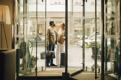 Couple with luggage talking while standing at doorway of hotel seen through glass