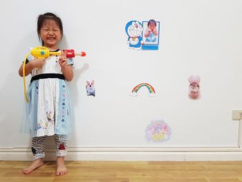 Smiling cute girl holding squirt gun while standing against wall at home