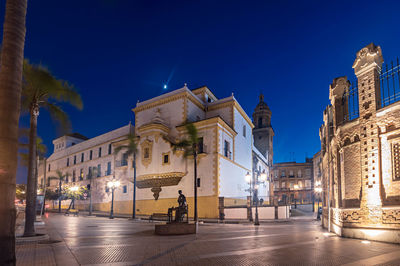 Illuminated street amidst buildings against blue sky at night