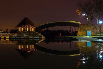 Illuminated bridge over river by buildings against sky at night