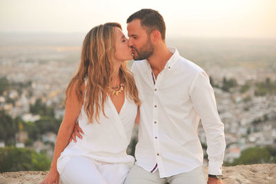 Young romantic couple kissing while sitting on retaining wall against cityscape