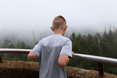 Rear view of teenage boy standing against forest during foggy weather