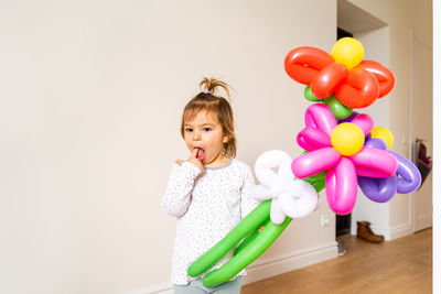 Cute girl with balloons standing against wall at home