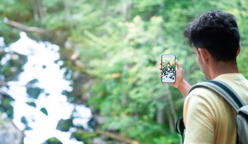 Back view of unrecognizable man traveler with backpack standing on slope taking picture with smartphone of amazing landscape of green forest with fast river