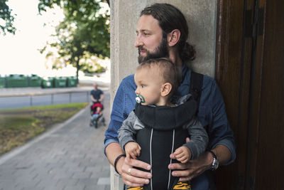 Thoughtful mid adult man carrying baby boy in carrier at doorway