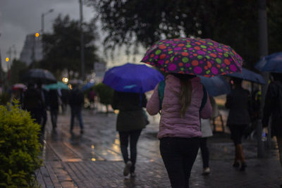 Rear view of people with umbrellas walking on wet footpath during rainy season at dusk