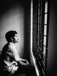 Side view of young man looking through window against wall at home