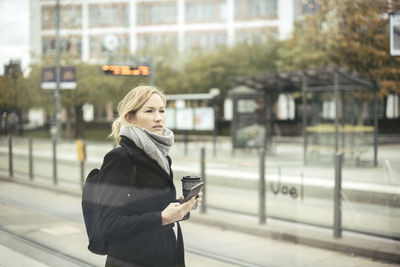 Mid adult businesswoman holding mobile phone and disposable coffee cup at tram station