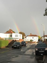 Cars on road by buildings in city against rainbow in sky