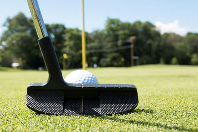 A putter is lined up behind a white golf ball with the yellow pin on the green in view