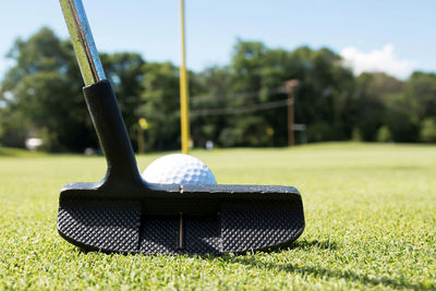 A putter is lined up behind a white golf ball with the yellow pin on the green in view