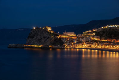 Illuminated buildings by sea against sky at night