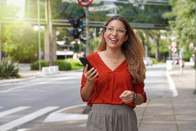 Smiling business woman holding mobile phone walking in modern sustainable metropolis