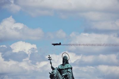 Low angle view of airplane flying over statue against cloudy sky