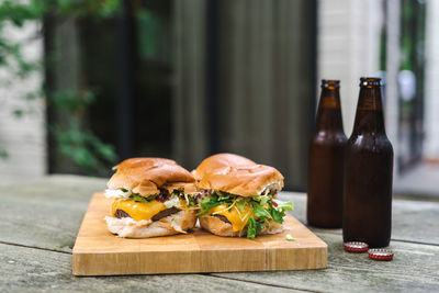 A set of cheeseburgers on a wooden plate and a couple of beers on an outdoor table. suburban.