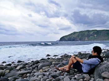 Man sitting on pebble stones at beach against sky