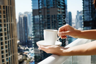 A girl holds a cup of coffee against the background of modern buildings. 