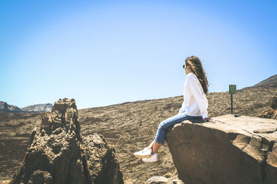 Side view of teenage girl looking at landscape while sitting on cliff against clear blue sky during sunny day