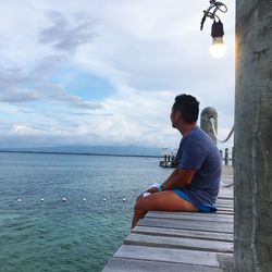 Side view of man looking at sea while sitting on pier against cloudy sky at lakawon