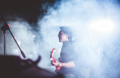 Man playing guitar while standing amidst smoke at nightclub