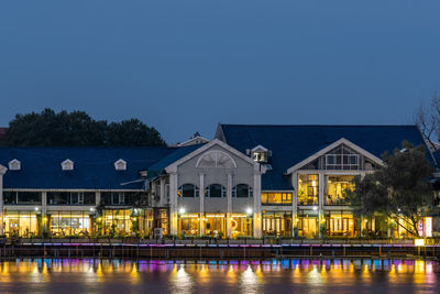 Illuminated building by river against sky at dusk