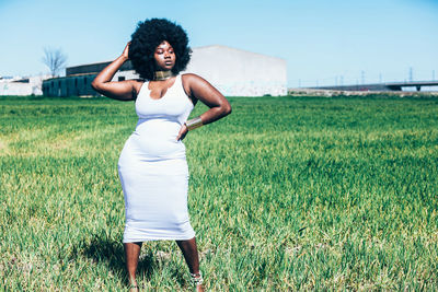 Full body curvy black female in trendy white dress with traditional african accessories and curly hair adjusting bracelet while standing on grass on cloudless day in field