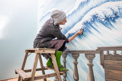Mature woman artist draws a mural on a marine theme sitting on a ladder