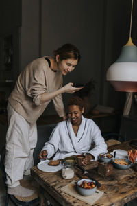 Non-binary person photographing friend having breakfast at home