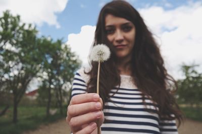 Portrait of smiling young woman showing dandelion against sky