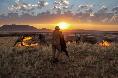 People extinguishing fire on field against sky during sunset