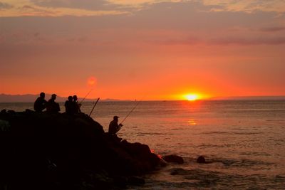 Silhouette friends fishing on sea shore against sky during sunset