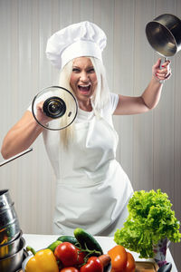 Portrait of young woman screaming while holding utensils in kitchen