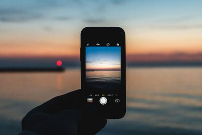 Person holding smart phone by sea against sky during sunset