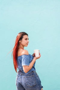 Portrait of young stylish woman with red hair drinking water against blue background