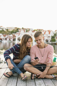 Full length of couple with fishing rod using mobile phone on wooden pier