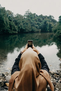 Rear view of woman photographing with smart phone by lake against trees and sky