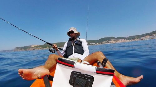 Man sitting on kayak while holding fishing rod at sea against clear blue sky