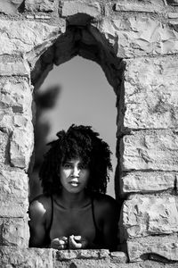 Portrait of young woman standing at window amidst stone wall