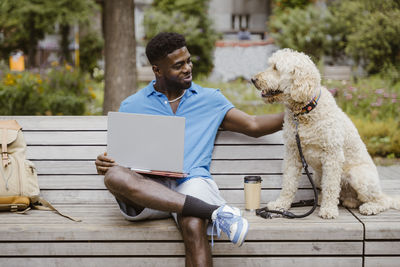 Smiling young man with laptop stroking dog while sitting on bench in park