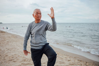 Senior man exercising while standing at beach against cloudy sky during sunset