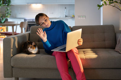 Young woman in blue sweater talks with friend via laptop videocall and shows domestic cat on sofa