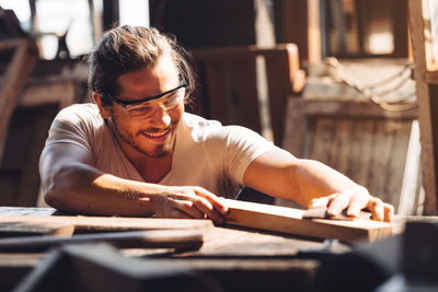 A young male carpenter builder working overall equals a wooden bar with a milling machine