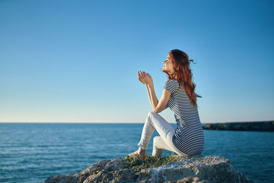 Side view of woman on rock by sea against blue sky